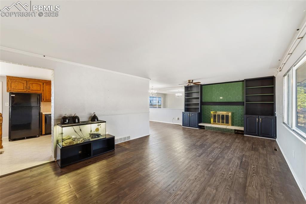 Living area featuring dark wood-type flooring, a brick fireplace, crown molding, built in shelves, and ceiling fan