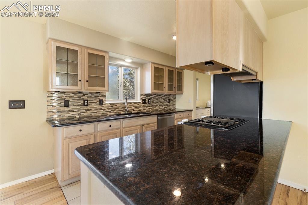 Kitchen with light brown cabinetry, dark stone counters, light wood-type flooring, glass insert cabinets, and backsplash