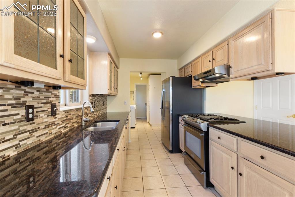 Kitchen with stainless steel range with gas stovetop, light brown cabinets, dark stone counters, glass insert cabinets, and light tile patterned floors