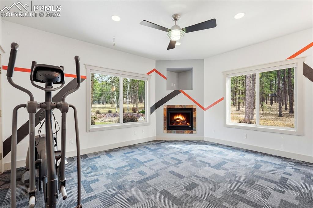 Exercise room with a warm lit fireplace, ceiling fan, light carpet / Was 2nd Shop Office