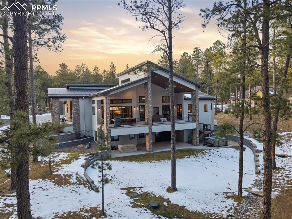 Snow covered property with a patio area, stucco siding, a metal roof, view of scattered trees, and stone siding