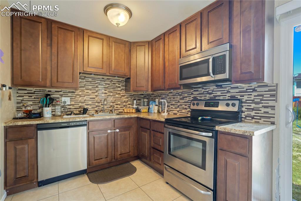 Kitchen featuring appliances with stainless steel finishes, light tile patterned floors, light stone countertops, and brown cabinetry
