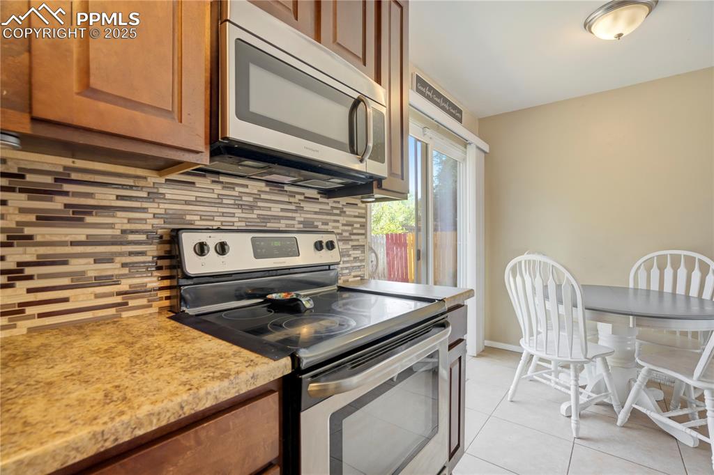 Kitchen with appliances with stainless steel finishes, brown cabinets, light tile patterned floors, light stone countertops, and tasteful backsplash