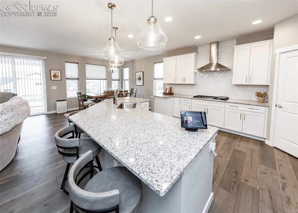 A beautifully designed kitchen with light wood-style flooring, elegant white cabinetry, and a decorative backsplash. The central island with sink is perfect for entertaining, complemented by a sleek wall-mounted chimney exhaust hood.