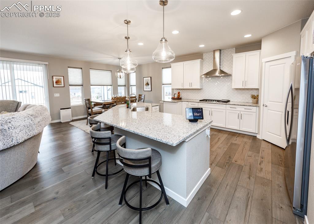 he kitchen features light wood-style flooring, white cabinetry, a decorative backsplash, and a functional island with sink. A wall-mounted chimney exhaust hood adds a modern finishing touch.