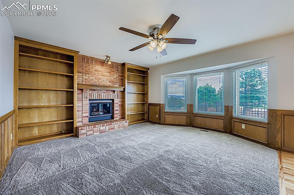 Unfurnished living room with built in features, wainscoting, ceiling fan, light carpet, and a brick fireplace