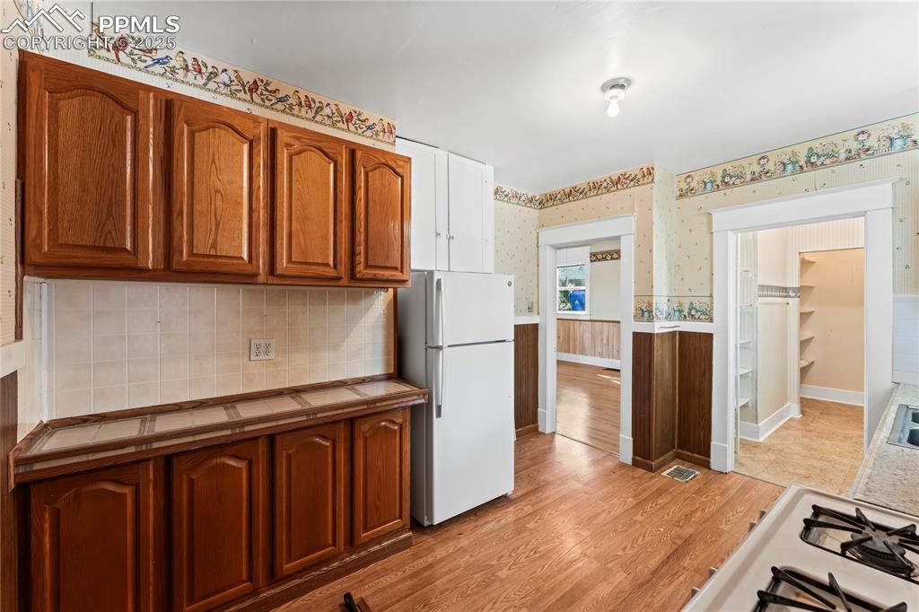 Kitchen featuring light wood-style flooring, brown cabinets, a wainscoted wall, white appliances, and wallpapered walls
