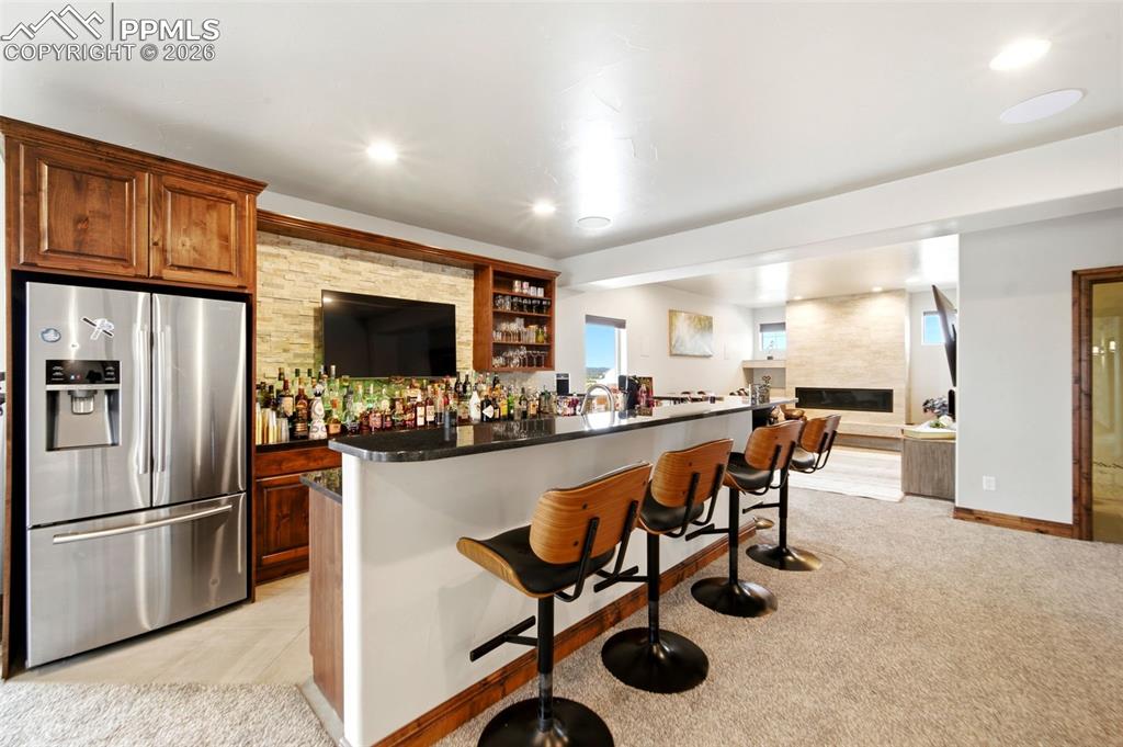 Indoor dry bar featuring stainless steel fridge with ice dispenser, light carpet, open shelves, a large fireplace, and recessed lighting