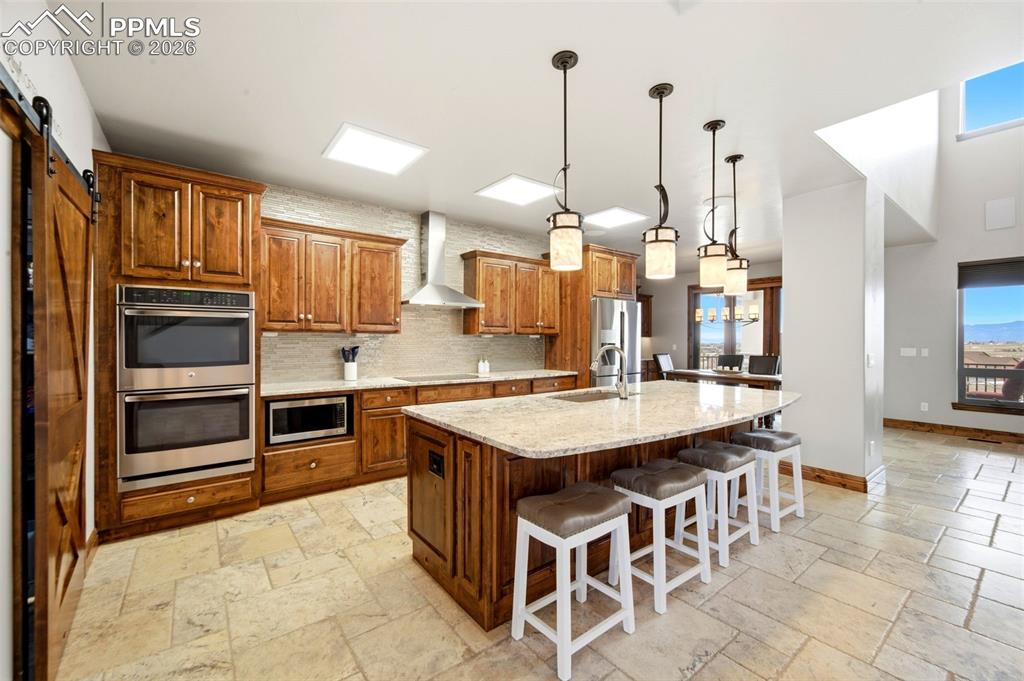 Kitchen featuring a barn door, wood finish cabinetry, stainless steel appliances, decorative light fixtures, and a center island with sink