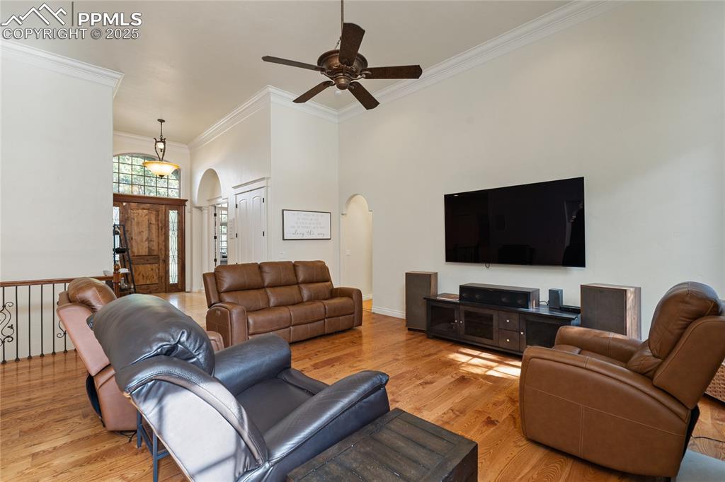 Living area featuring a towering ceiling, light wood-style flooring, a ceiling fan, crown molding, and arched walkways
