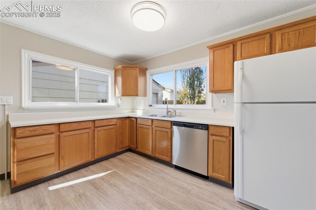 Kitchen featuring freestanding refrigerator, stainless steel dishwasher, light countertops, and brown cabinetry