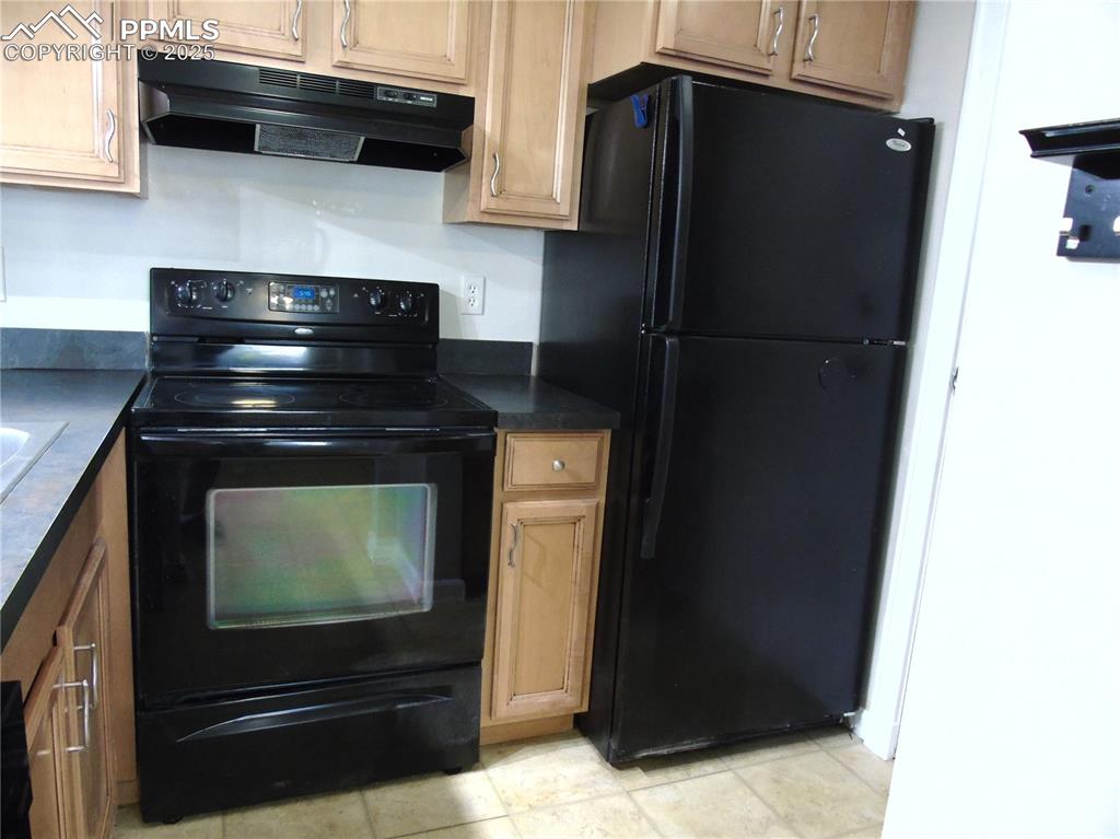 Kitchen with black appliances, dark countertops, under cabinet range hood, and light tile patterned floors