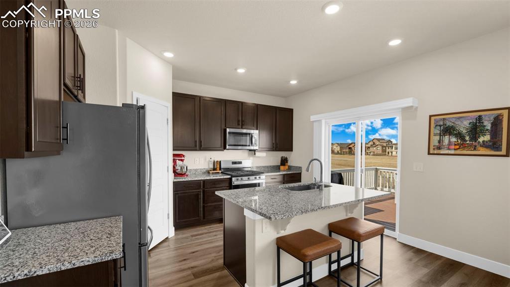 Kitchen with dark wood finish cabinets, light stone countertops, light wood-style flooring, stainless steel appliances, and a kitchen breakfast bar