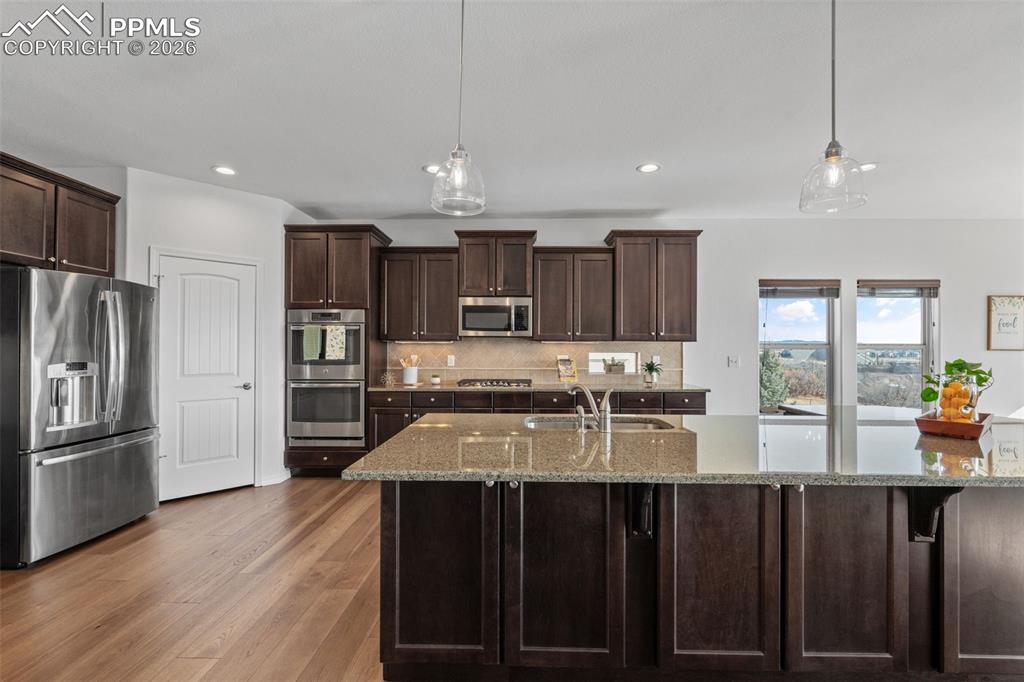 Kitchen with dark wood finish cabinetry, stainless steel appliances, light stone countertops, and pendant lighting