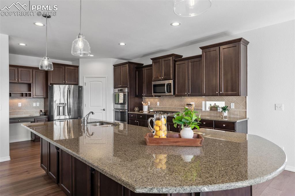 Kitchen featuring dark wood finish cabinetry, dark wood-type flooring, stainless steel appliances, hanging light fixtures, and dark stone countertops