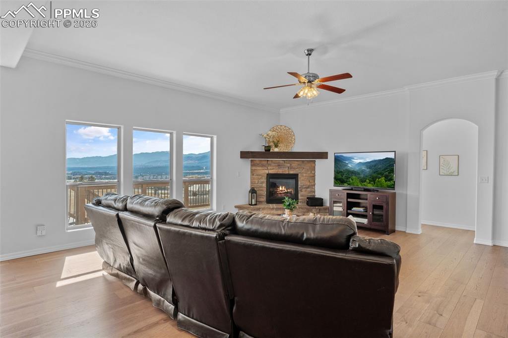 Living room featuring crown molding, arched walkways, light wood finished floors, ceiling fan, and a stone fireplace