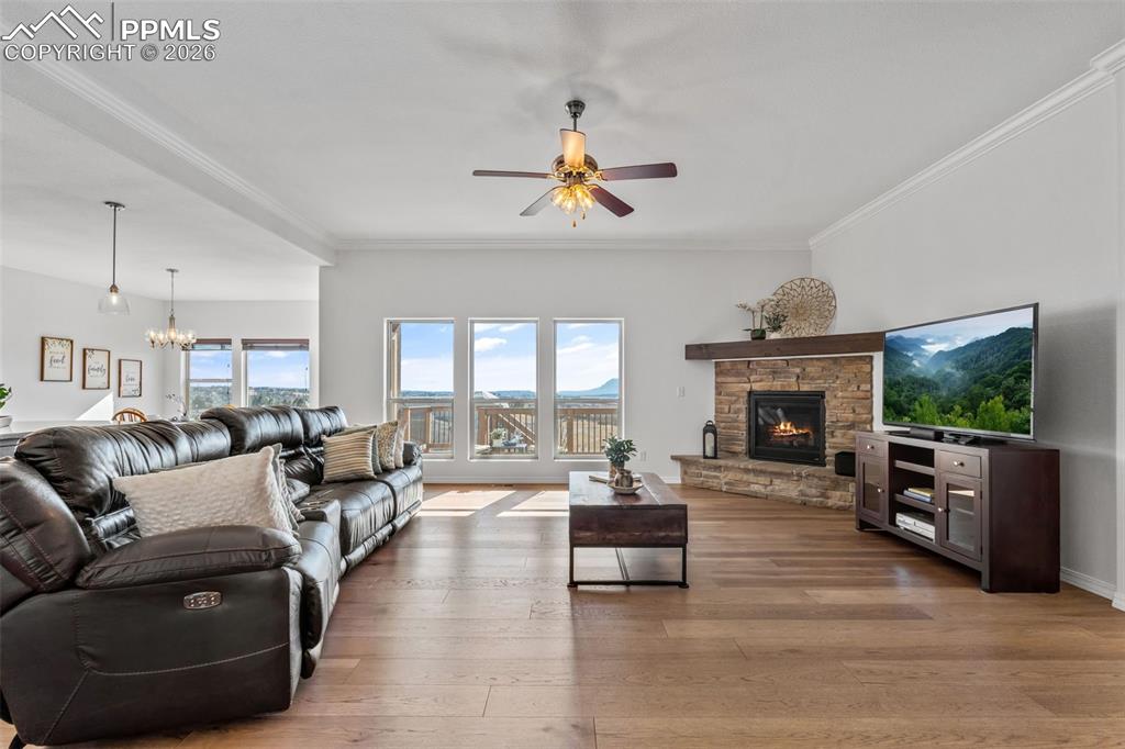 Living area featuring crown molding, a stone fireplace, light wood-style flooring, a ceiling fan, and suspended lighting