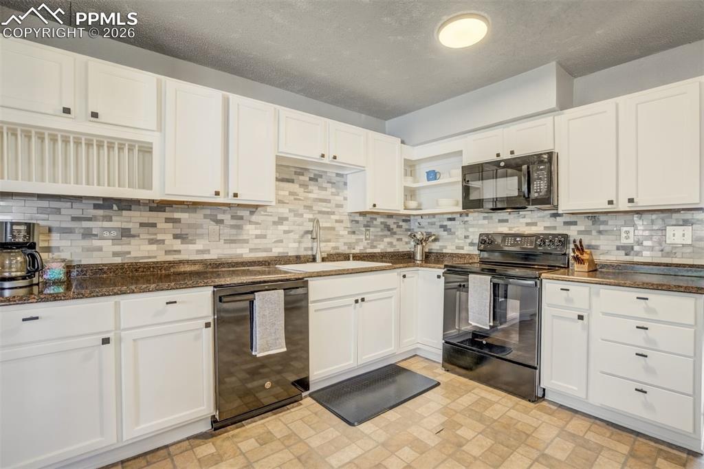 Kitchen featuring black appliances, white cabinetry, open shelves, dark stone counters, and brick patterned floors