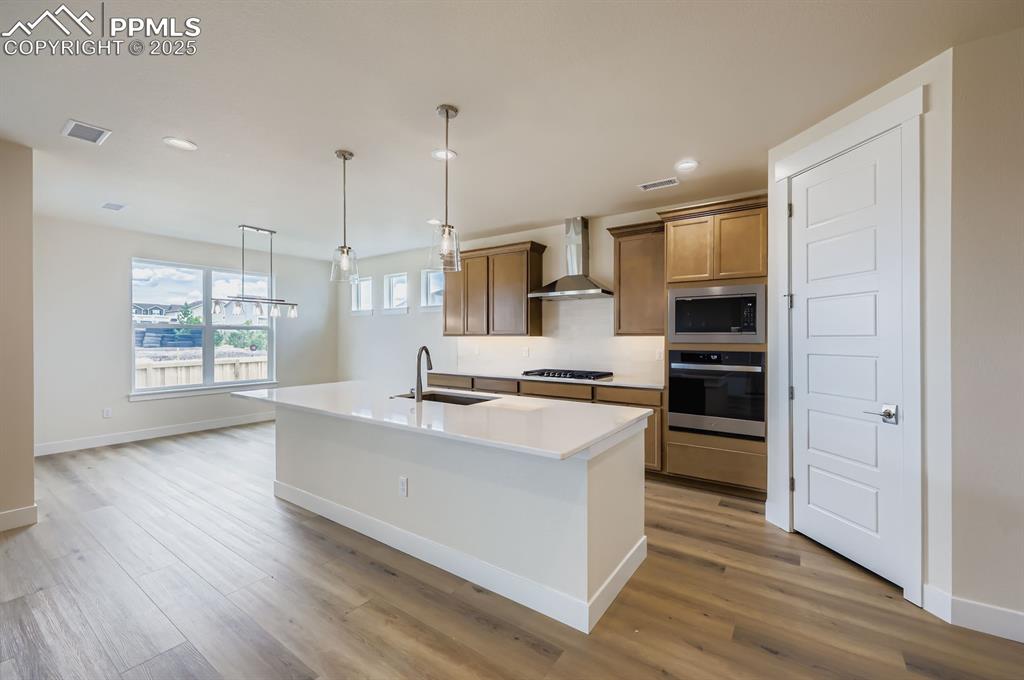Kitchen with stainless steel appliances, wall chimney range hood, light wood finished floors, recessed lighting, and a center island with sink