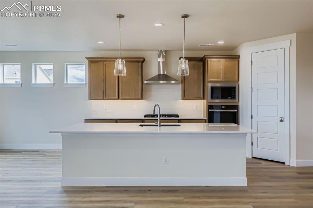 Kitchen featuring wall chimney exhaust hood, stainless steel appliances, decorative light fixtures, light countertops, and a center island with sink