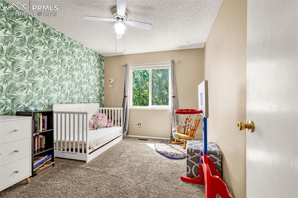 Bedroom with carpet, wallpapered walls, a nursery area, ceiling fan, and a textured ceiling