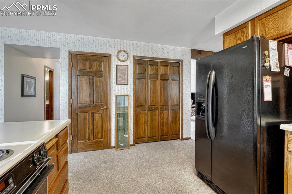 Kitchen featuring black refrigerator with ice dispenser, light colored carpet, brown cabinetry, stainless steel range oven, and light countertops