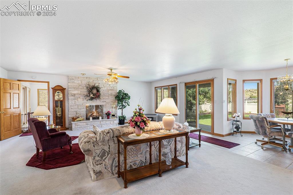Carpeted living room featuring a stone fireplace and ceiling fan with notable chandelier