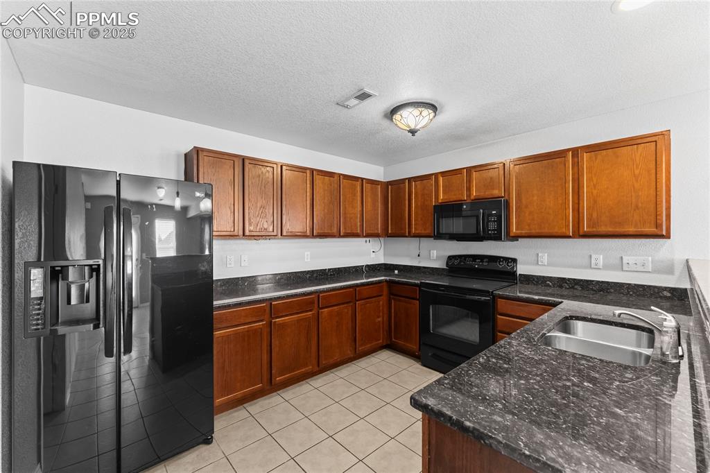 Kitchen featuring black appliances, light tile patterned floors, dark stone countertops, brown cabinetry, and a textured ceiling