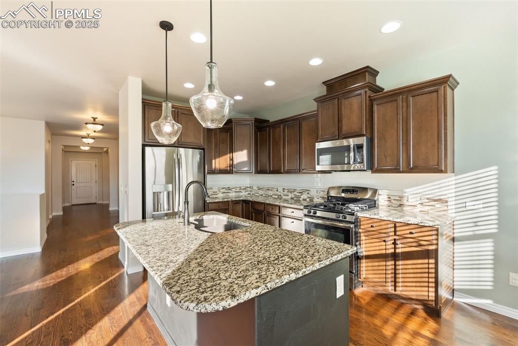 Kitchen with appliances with stainless steel finishes, light stone countertops, decorative light fixtures, dark brown cabinets, and a center island with sink