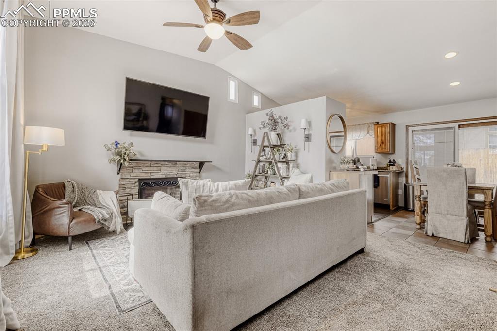 Living room featuring ceiling fan, vaulted ceiling, a stone fireplace, and light colored carpet