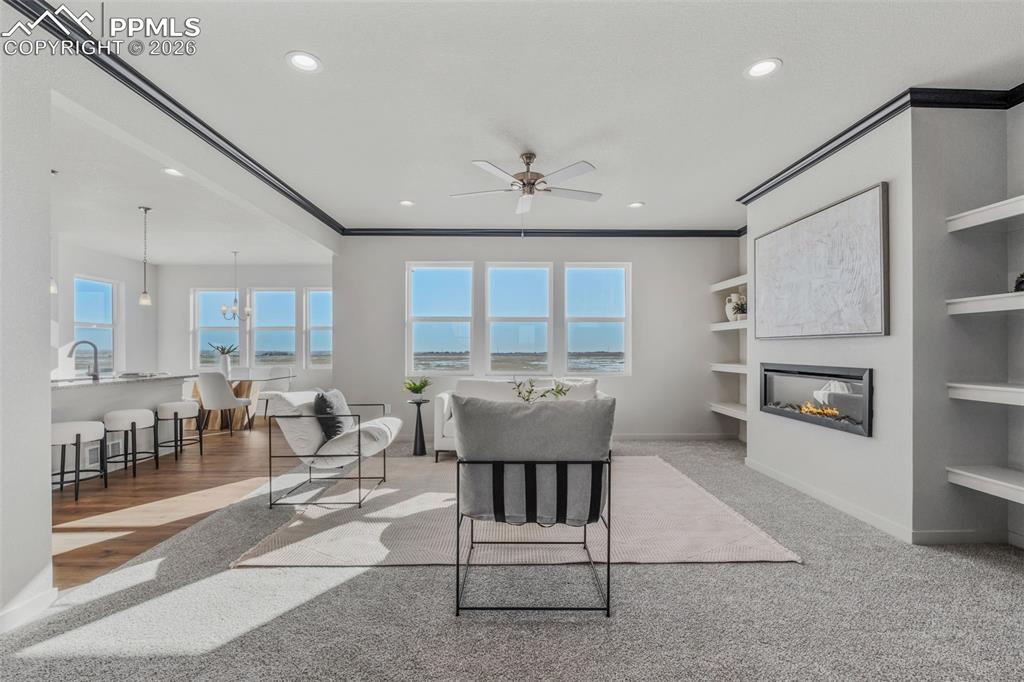 Living room with built in features, a glass covered fireplace, ceiling fan, crown molding, and healthy amount of natural light