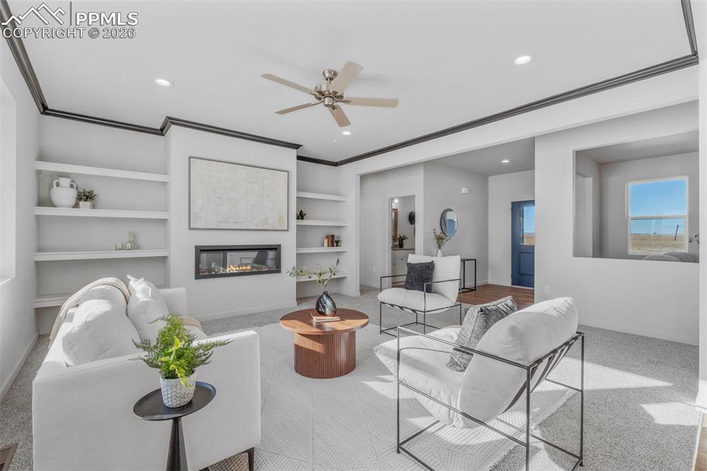 Living room with built in shelves, a glass covered fireplace, carpet floors, ceiling fan, and ornamental molding
