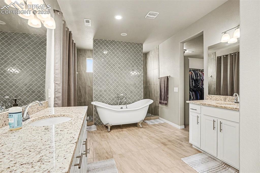 Bathroom featuring two vanities, a walk in closet, a soaking tub, tile walls, and recessed lighting
