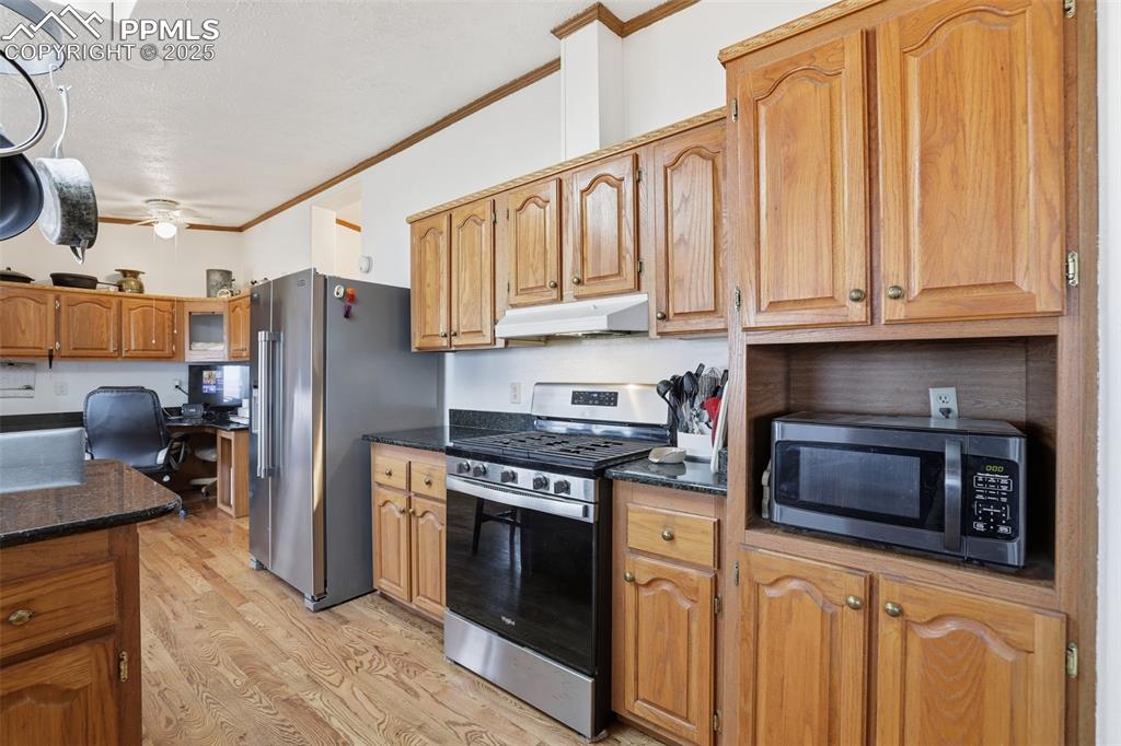 Kitchen with ornamental molding, stainless steel appliances, light wood-style flooring, dark stone counters, and under cabinet range hood