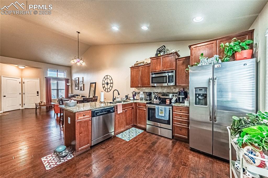 Kitchen with stainless steel appliances, lofted ceiling, light stone countertops, hanging light fixtures, and dark wood-style floors