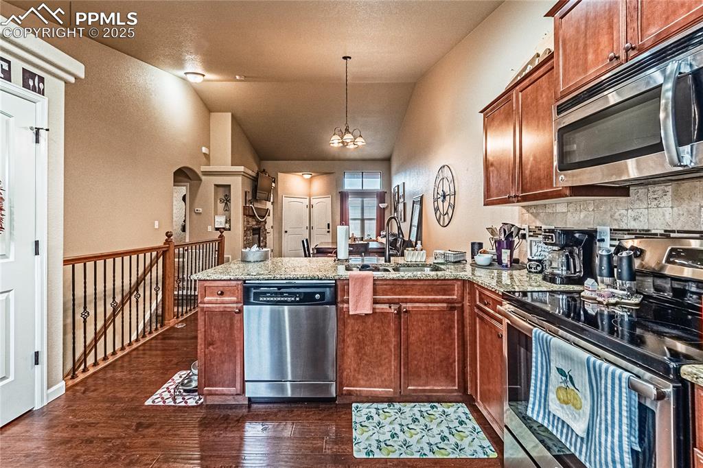 Kitchen with appliances with stainless steel finishes, a peninsula, brown cabinetry, vaulted ceiling, and dark wood-type flooring