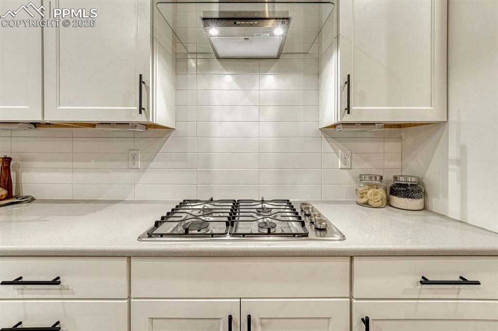 Kitchen equipped with ventilation hood, white cabinetry, stainless steel gas cooktop, and decorative backsplash.