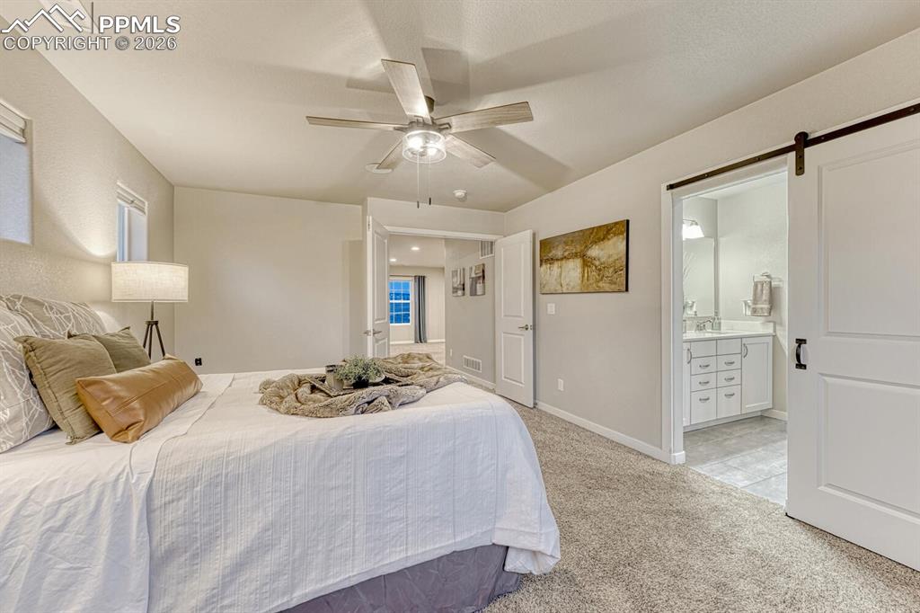 Bedroom with a barn door, light colored carpet, ceiling fan, and ensuite bath