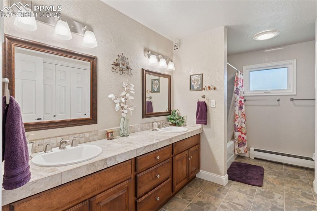 Full bathroom with double vanity, shower / bath combo, a textured wall, and stone flooring that connects multiple ground level bedrooms