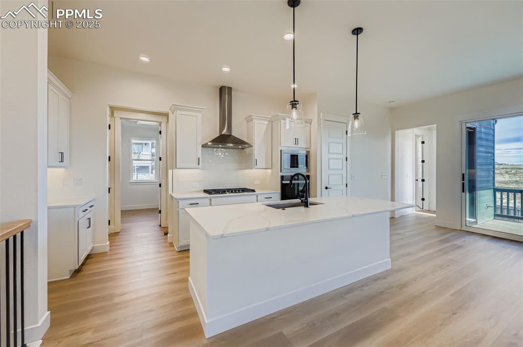 Kitchen with white cabinets, wall chimney range hood, an island with sink, decorative light fixtures, and recessed lighting