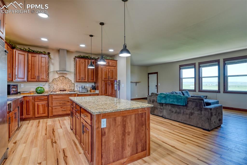 Kitchen featuring wall chimney range hood, brown cabinets, light wood-style floors, a kitchen island, and light stone counters