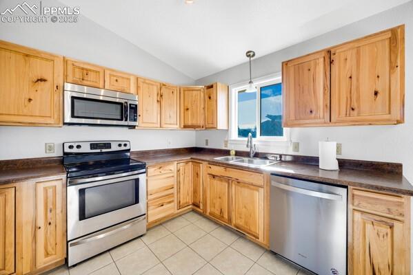 Kitchen with appliances with stainless steel finishes, dark countertops, decorative light fixtures, light brown cabinetry, and vaulted ceiling