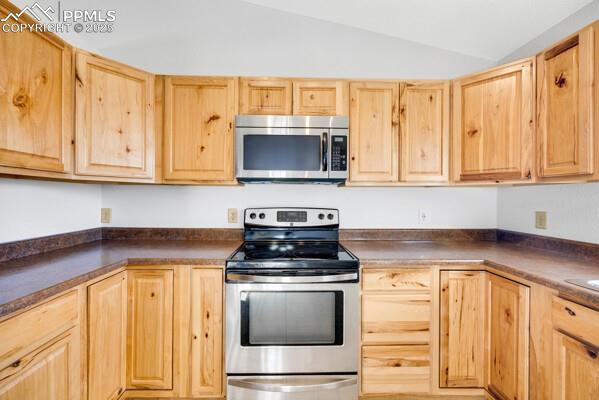 Kitchen featuring stainless steel appliances, light brown cabinets, vaulted ceiling, and dark countertops