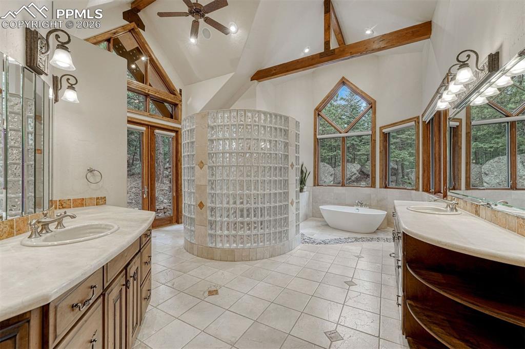 Vaulted ceilings and exposed beams frame a spa-like primary bathroom with dual vanities, soaking tub, and serene nature views.