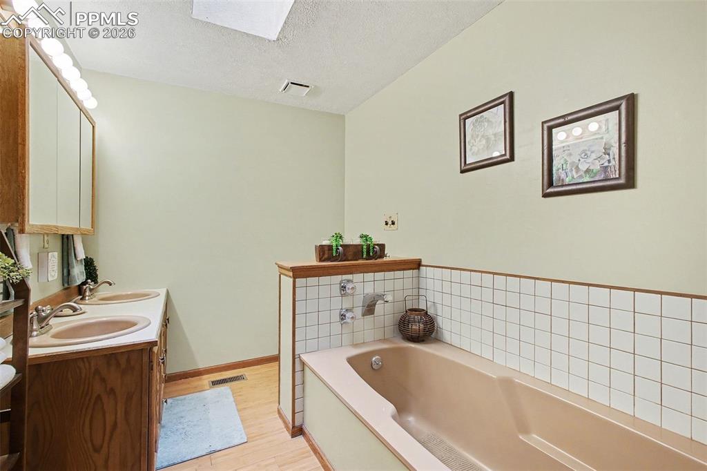 Bathroom featuring double vanity, a bath, a textured ceiling, light wood finished floors, and a skylight