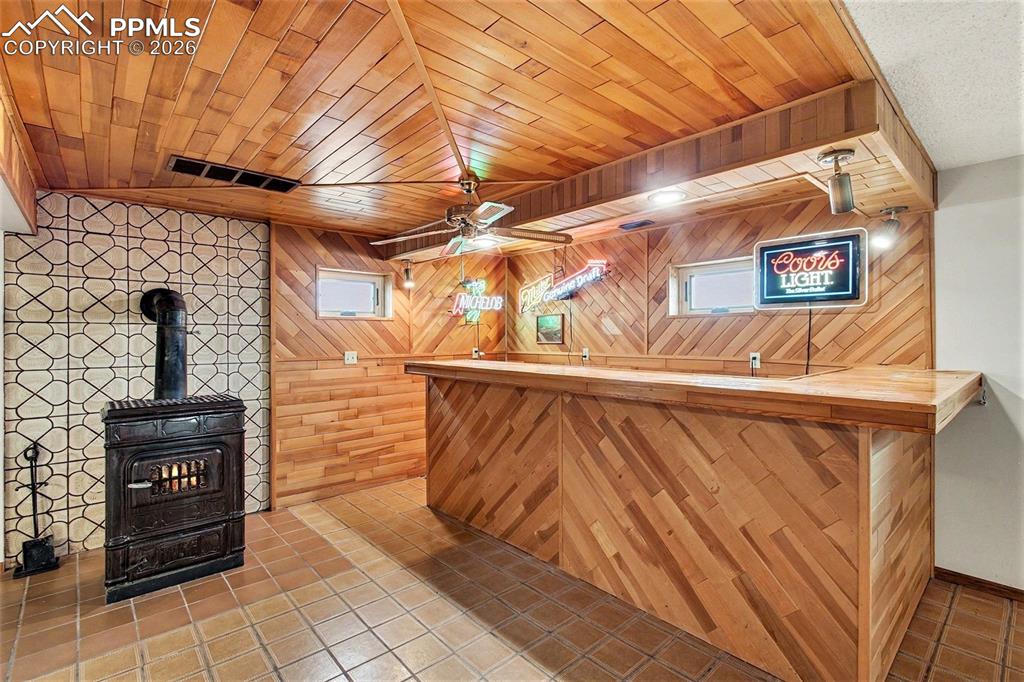 Indoor dry bar with a wood stove, wood walls, tile patterned floors, wood ceiling, and ceiling fan