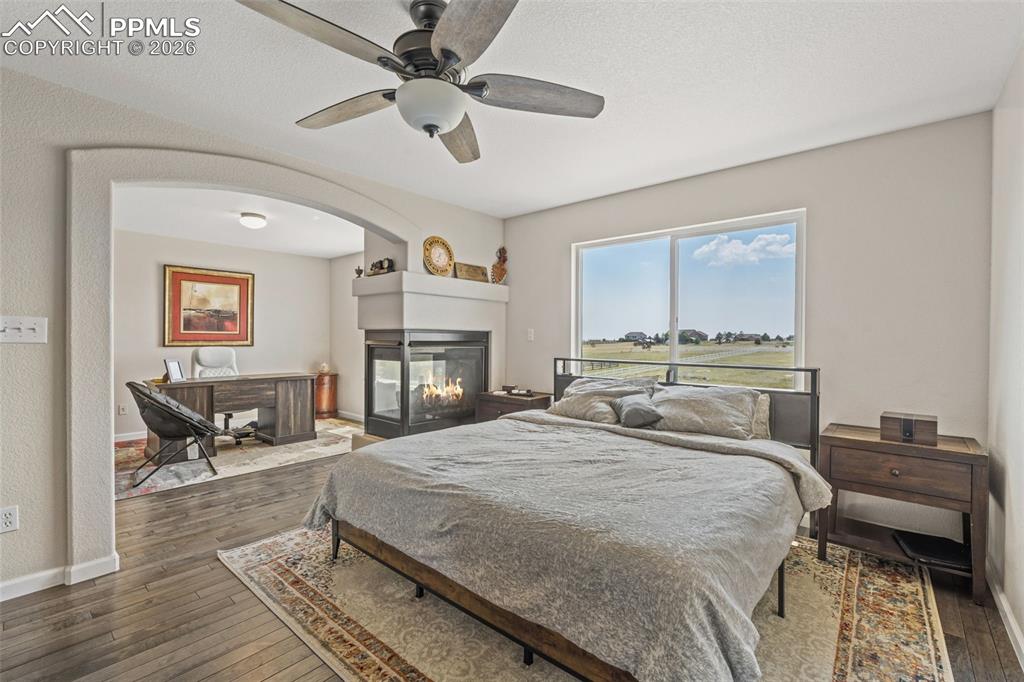 Bedroom featuring dark wood-style flooring, a ceiling fan, a desk, and a multi sided fireplace