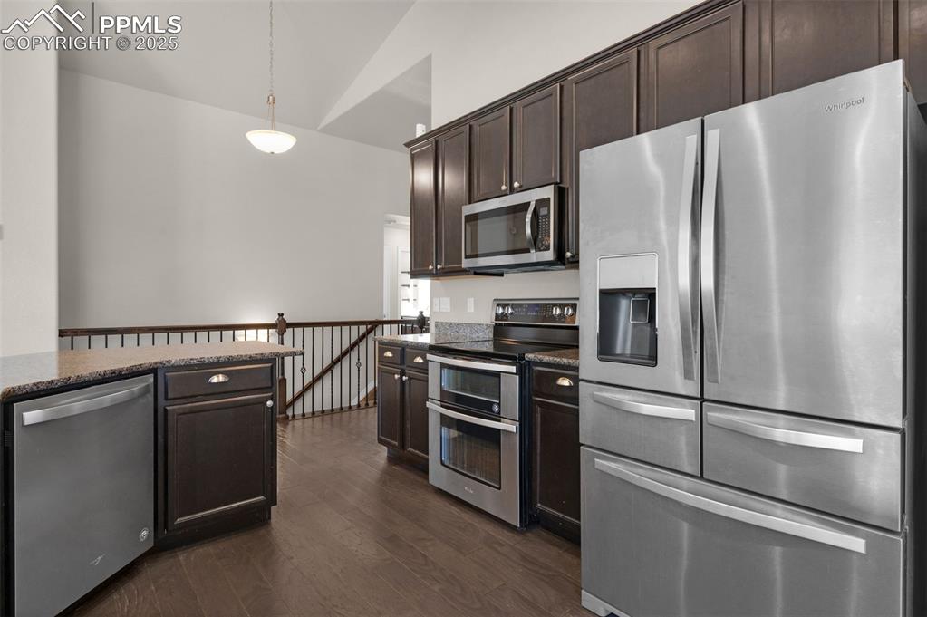 Kitchen with appliances with stainless steel finishes, decorative light fixtures, dark wood-type flooring, dark stone countertops, and lofted ceiling