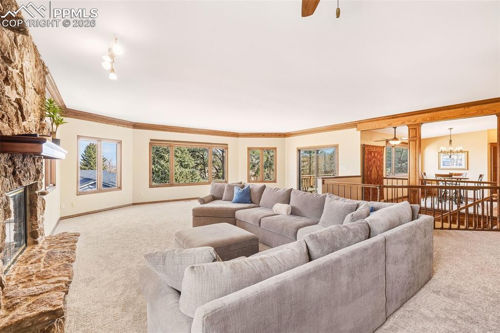 Expansive main-level living area with plush carpet, crown molding, and a striking stone fireplace that anchors the space. Large windows frame treetop views to Downtown Colorado Springs, while the open layout flows seamlessly to the dining area and deck access.