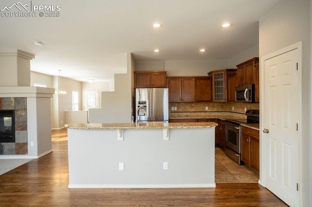 Kitchen with stainless steel appliances, glass insert cabinets, light stone counters, a kitchen island with sink, and dark wood-style flooring